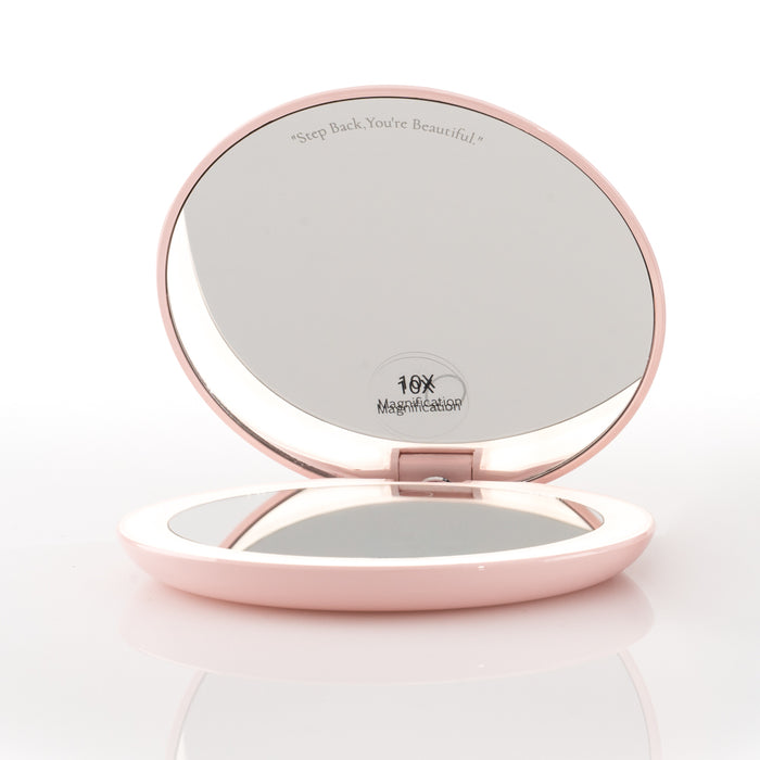 The open Nikol Beauty 10x Magnification Mirror compact in pink reveals two mirrors. The top features the engraved phrase "Step Back, You’re Beautiful" above the 10X magnification label, resting on a white surface.