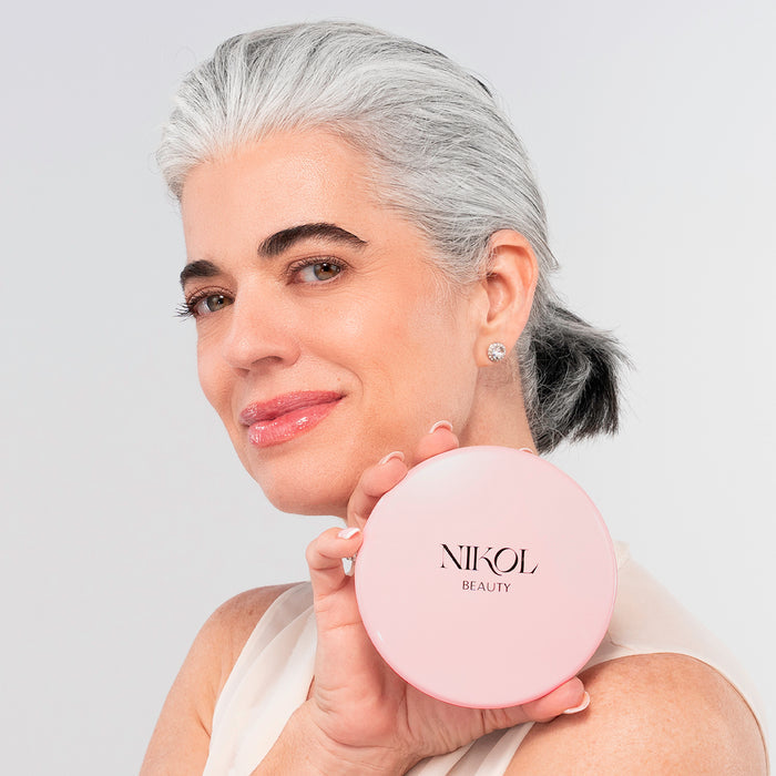 A woman with gray hair pulled back smiles and holds the cordless Nikol Beauty 10x Magnification Mirror near her face, as if admiring herself. She wears a sleeveless top and stud earrings, set against a light background.