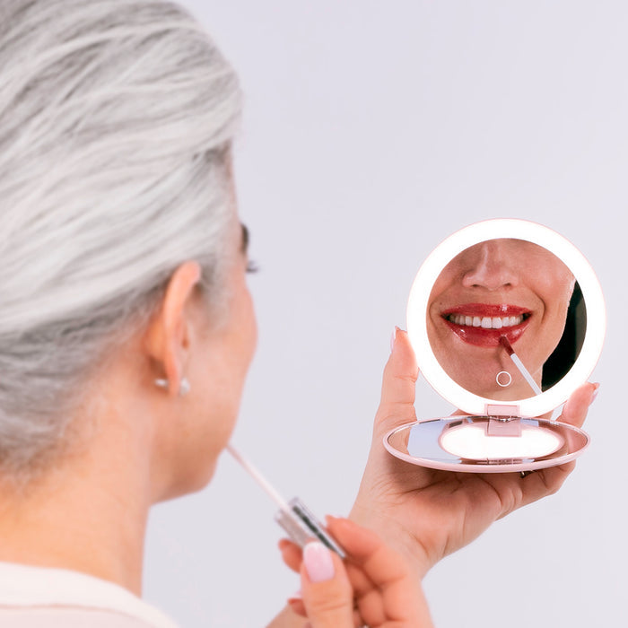 A woman with gray hair applies lip gloss while looking into the round, illuminated Nikol Beauty 10x Magnification Mirror, which reflects her smiling lips.