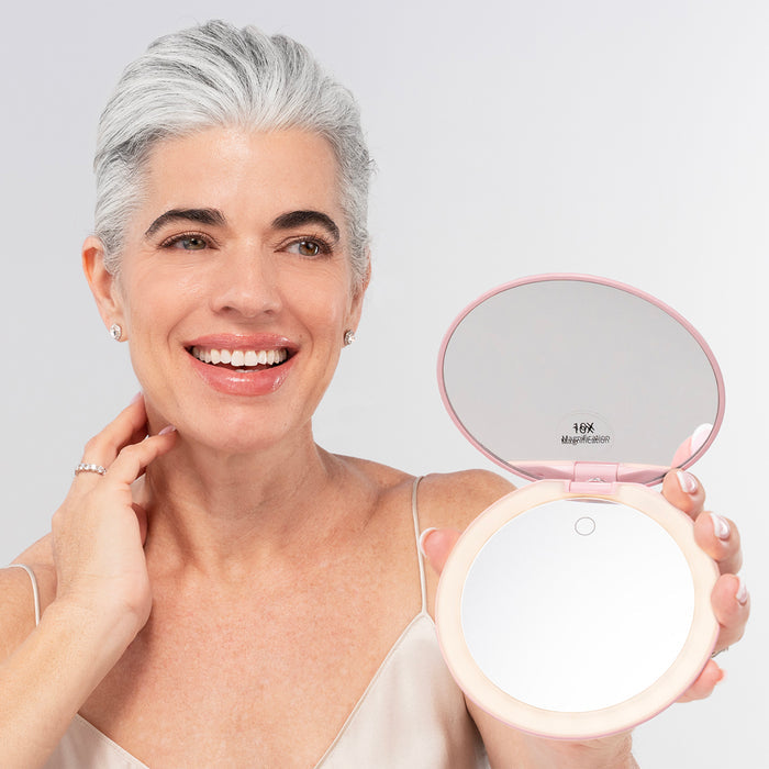 A smiling woman with short gray hair holds the cordless Nikol Beauty 10x Magnification Mirror in one hand, a rechargeable LED ring light makeup mirror with no cable attached. She wears a beige camisole and small earrings against a neutral background.