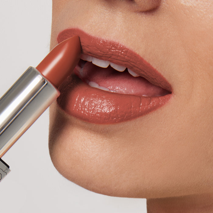 A close-up shows someone applying Nikol Beautys 1975 Shiny Velvet Cream Lipstick #9, an ultra-creamy, brownish-red shade. It glides smoothly from the metallic tube onto slightly open lips, providing a glossy finish against a neutral background.
