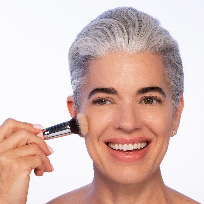 A smiling woman with short gray hair applies Nikol Beauty BB CRÈME to her cheek using a brush. She has light, mature skin, wears stud earrings, and is posed against a plain white background.