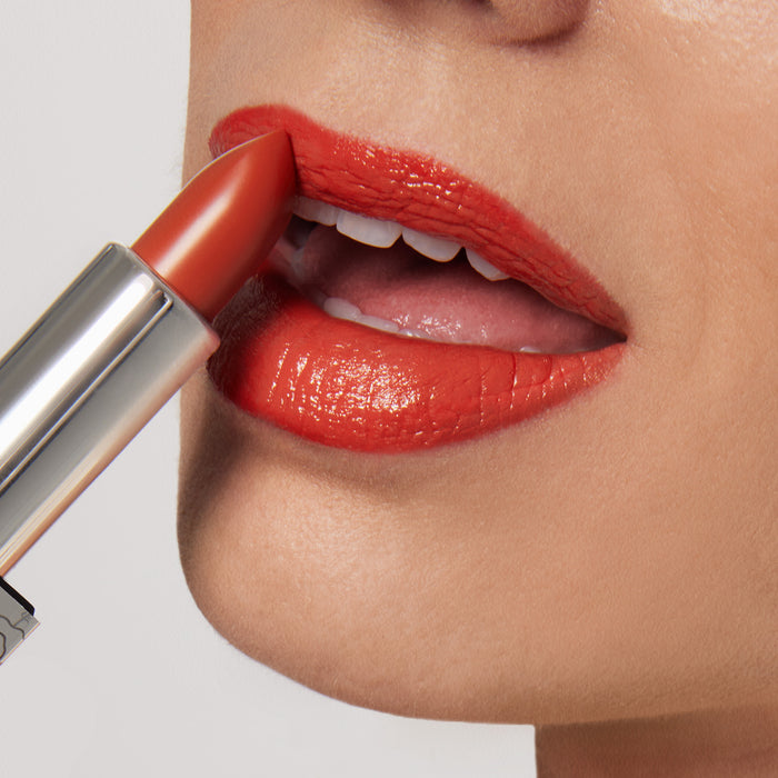 A close-up captures a person applying Nikol Beautys Beach House Shiny Velvet Cream Lipstick #122, in a bright orange hue with hydrating shea butter. Their parted lips reveal teeth, showcasing the vegan lipstick’s shiny finish against a soft, light background.