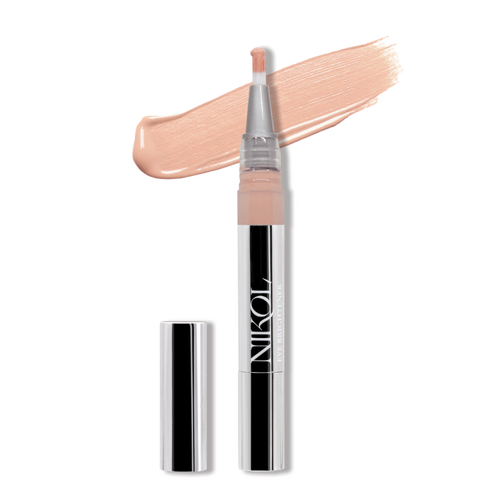 The Eye Brightener pen by Nikol Beauty, uncapped and vertical, showcases a clear and silver design with an eye brightener formula. Nearby is a light pink concealer swatch. The pen, enhanced with Chamomile Flower Water, prominently displays the brand name on its body.