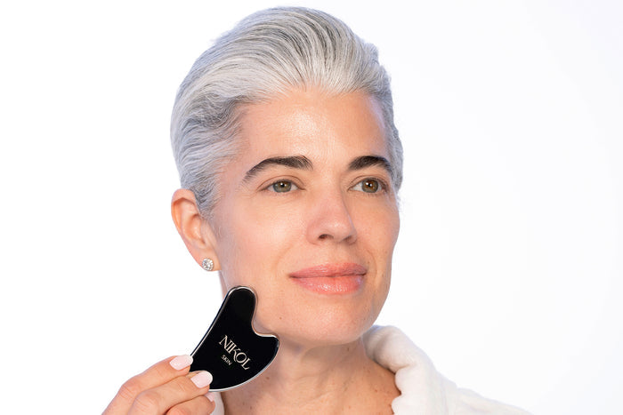 A woman with short gray hair smiles softly as she uses the Nikol Beauty Gua Sha Stainless Steel tool on her cheek, promoting lymphatic drainage. She wears a white top and stud earrings against a light background.
