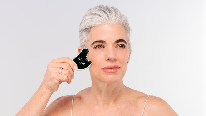 A woman with short gray hair uses the Nikol Beauty Gua Sha Stainless Steel facial massage tool on her cheek. She wears a light camisole and appears calm, set against a plain, light background.