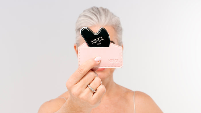 A woman with short gray hair holds a black Gua Sha Stainless Steel tool from Nikol Beauty in a pale pink case, partially covering her face. She wears a thin strap top and silver ring, set against a plain white background.