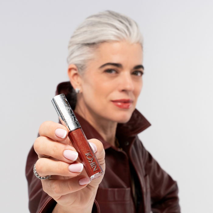 A mature woman with short gray hair and a maroon jacket confidently holds up Nikol Beauty Ignite Lip Gloss #176 toward the camera, her face softly blurred in the background as the lip gloss is in sharp focus.