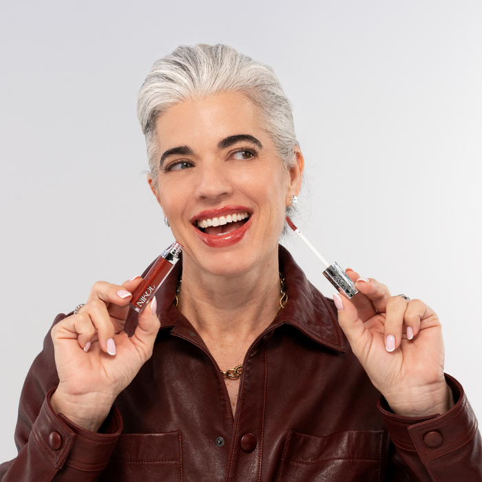 A smiling mature woman with short gray hair in a maroon leather jacket holds Nikol Beauty’s Ignite Lip Gloss #176, showing off her freshly applied high-shine red lips against a plain light background.