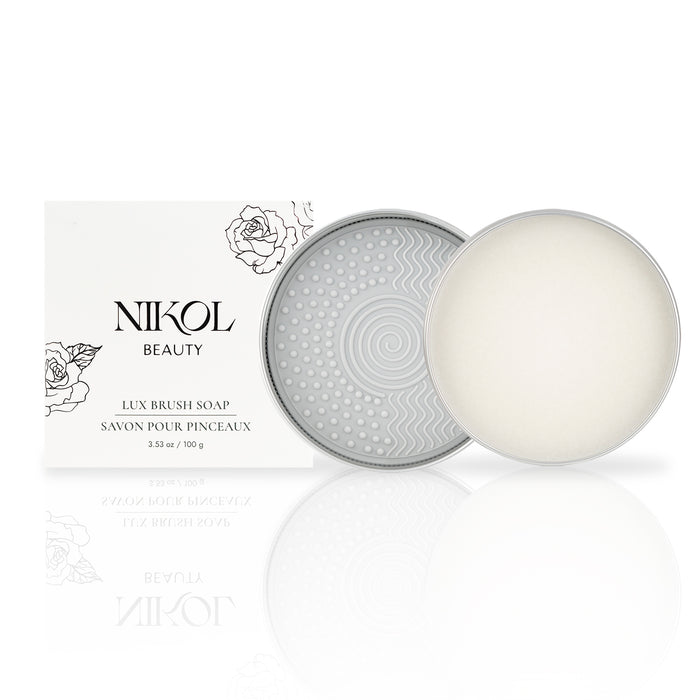 A cylindrical tin with a textured geometric lid holds a round bar of Lux Brush Soap by Nikol Beauty next to its white packaging adorned with floral line art. Infused with coconut oil, the soap gently cleans makeup brushes.
