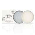 A cylindrical tin with a textured geometric lid holds a round bar of Lux Brush Soap by Nikol Beauty next to its white packaging adorned with floral line art. Infused with coconut oil, the soap gently cleans makeup brushes.