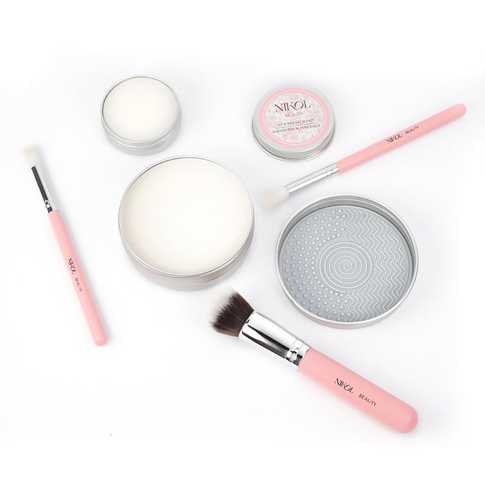 A flat lay of Nikol Beauty Lux Brush Soap, pink-handled brushes, tins of vegan brush cleaner, coconut oil brush soap, and a round cleaning pad, all neatly arranged on a white background.