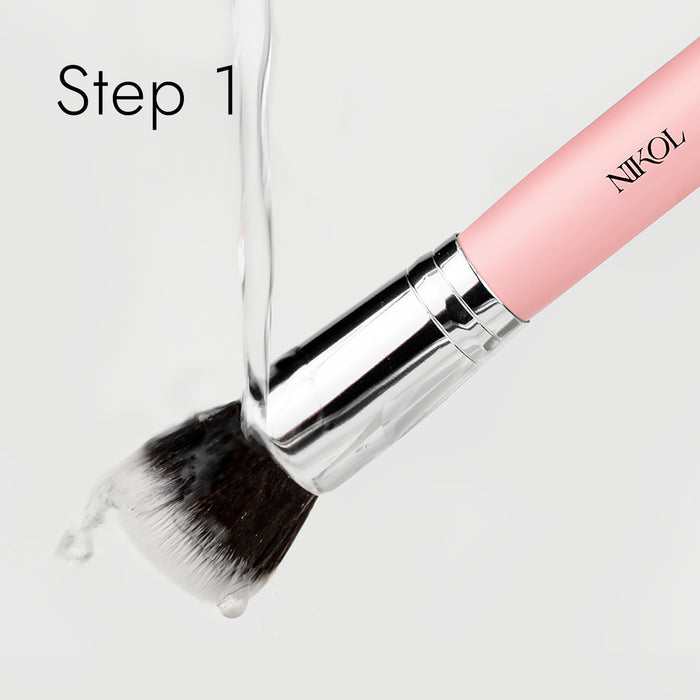 A pink-handle makeup brush with a silver ferrule is washed under running water, its bristles glistening wet. The process begins with Nikol Beautys Lux Brush Soap enriched with coconut oil. Step 1 is displayed in the top left corner.