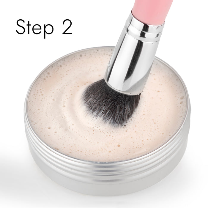 A makeup brush swirls in a round container filled with light-colored soapy water, revitalized by Nikol Beautys Lux Brush Soap. The words Step 2 adorn the upper left corner.