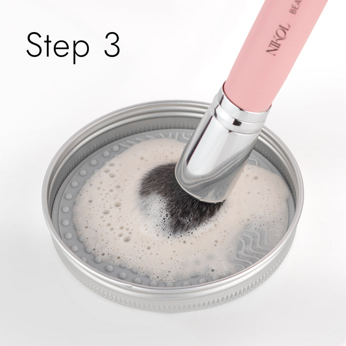A makeup brush with a pink handle and silver ferrule is cleaned in a lid filled with Nikol Beautys Lux Brush Soap infused with soapy water. Bubbles dance on the surface, while Step 3 in the top left corner ensures your beauty routine stays pristine.