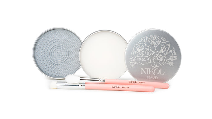 Two round silver tins—one with a textured pad and the other with Lux Brush Soap by Nikol Beauty—are displayed open. In front, two pink-handled Nikol Beauty makeup brushes rest on a white background.