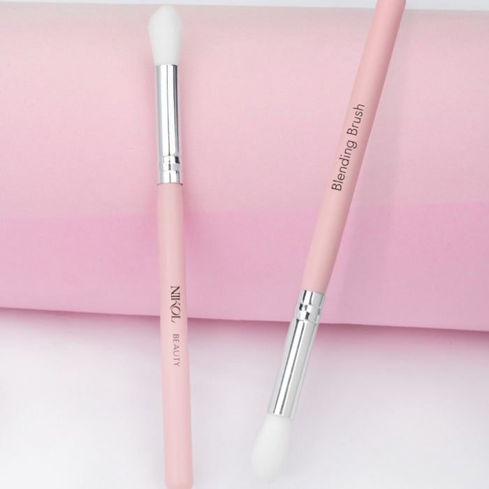 On a pink and white gradient background are two pink makeup brushes with white bristles. One handle reads Nikol Beauty, while the other, labeled Blending Brush, is ideal for mature eyes. Featuring silver ferrules, both are crafted from 100% vegan materials by Nikol Beauty.