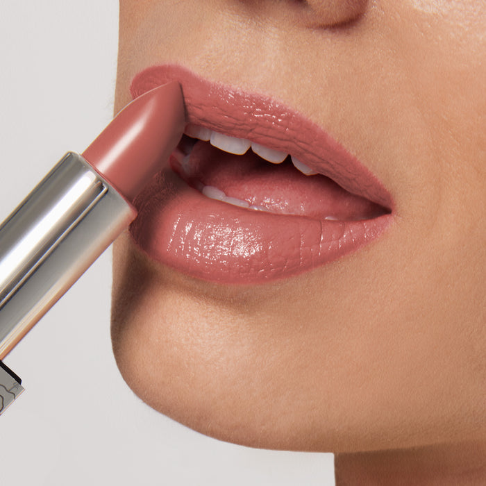 A person applies NikRay Shiny Velvet Cream Lipstick #701 from Nikol Beauty, a nude pink hydrating lipstick in a silver tube praised for its cruelty-free formula. Focus on their glossy, mature lips and chin as the partially extended lipstick nears them.