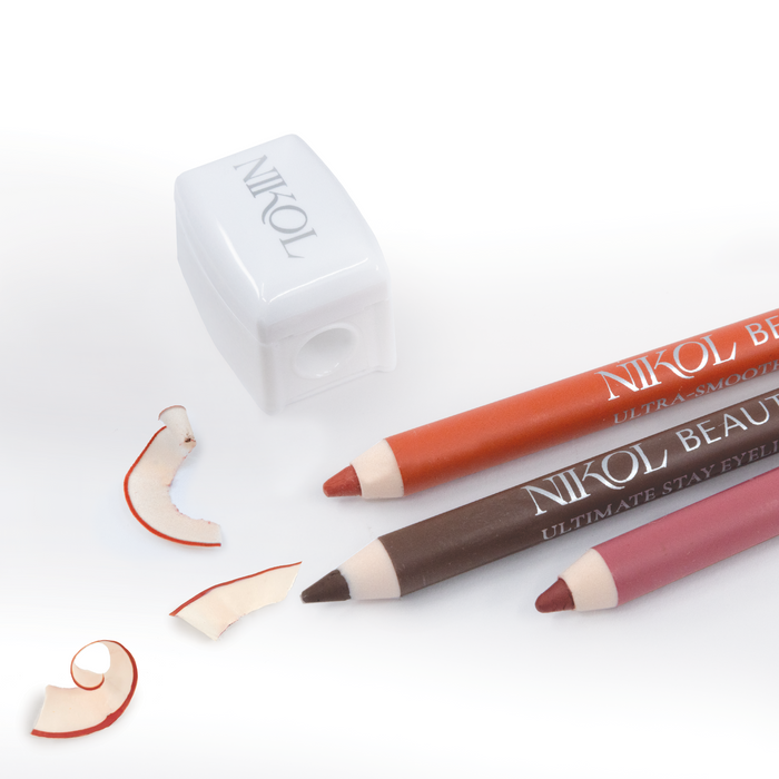 Two Nikol Beauty eyeliners, in brown and red, are elegantly placed beside a Pencil Sharpener on a white surface. Scattered around are shavings that have red-tinged edges, showcasing professional application—a true makeup bag essential.
