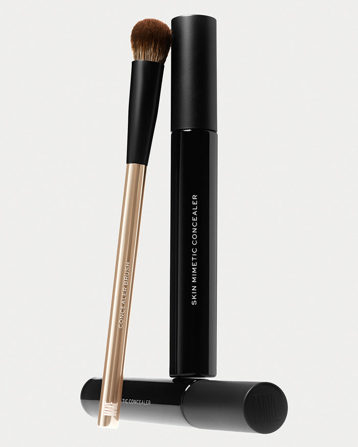 Skin Mimetic Concealer tube in sleek black packaging, shown with its cap removed and paired with a Concealer Brush featuring a fluffy tip and gold handle ideal for seamless, buildable concealer application.