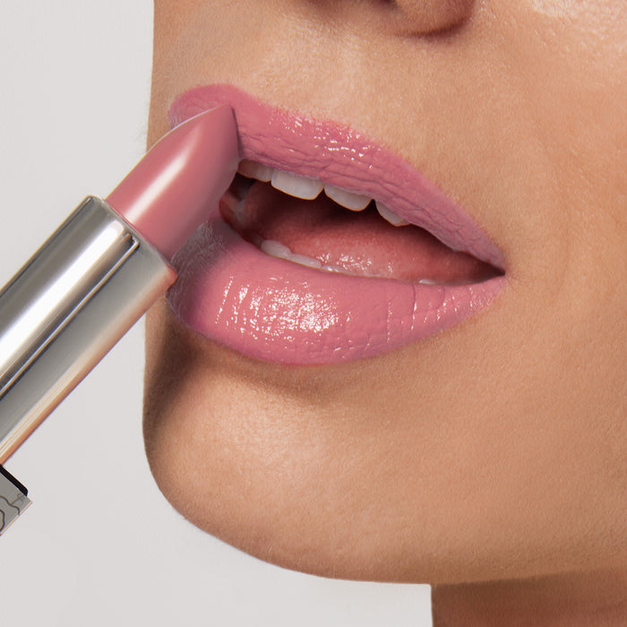 A person applies Nikol Beautys Pretty Smart Shiny Velvet Cream Lipstick #337, showcasing its ultra-creamy pink finish. The metallic reflective case suggests its hydrating formula, with slightly parted lips revealing a hint of white teeth against a neutral gray background.