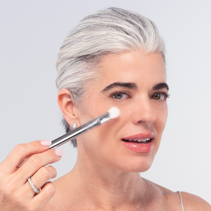 A woman with short gray hair applies Nikol Beauty’s SOFT SET TECHNIQUE™ BRUSH to her cheek. She wears a light top, stud earrings, and a ring, smiling softly against a plain light background.