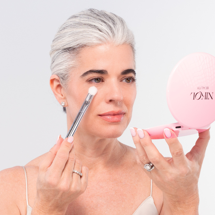 A woman with gray hair applies makeup using the Nikol Beauty SOFT SET TECHNIQUE™ BRUSH while holding a pink compact mirror. Wearing a light-colored top and rings, she looks focused on her reflection.