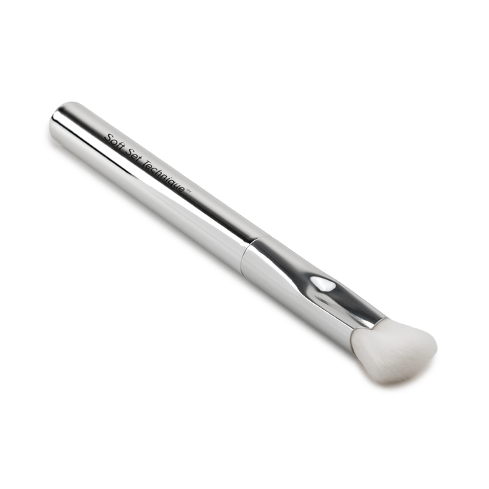 The SOFT SET TECHNIQUE™ BRUSH by Nikol Beauty is a silver vegan makeup brush with a sleek metallic handle and soft, angled white bristles—perfect as an under-eye setting brush for mature skin. Shown on a white background.