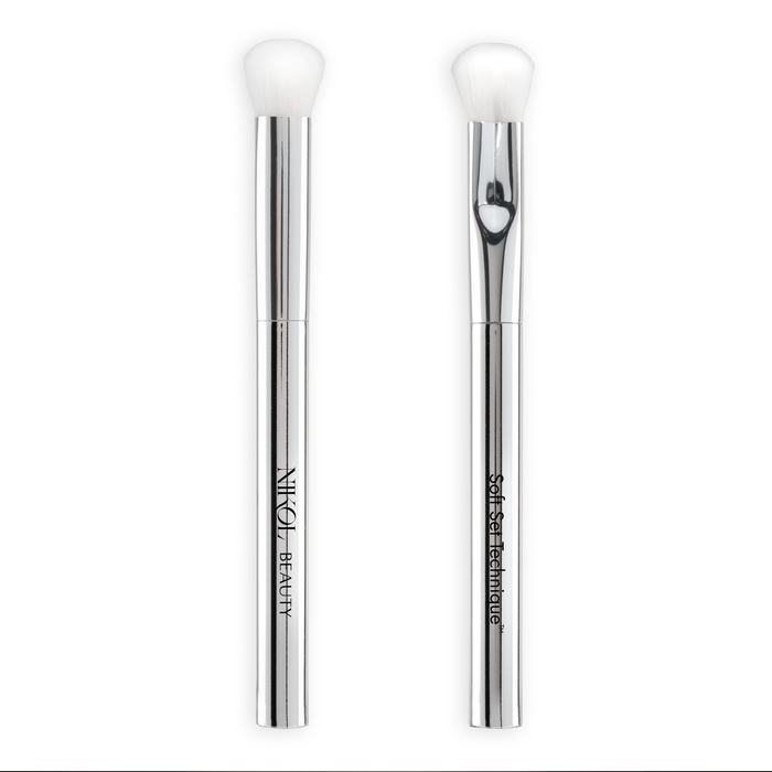 Two silver-handled vegan makeup brushes with white bristles stand upright against a white background. One handle is labeled Nikol Beauty and the other SOFT SET TECHNIQUE™ BRUSH—ideal for mature skin or setting under-eye makeup.