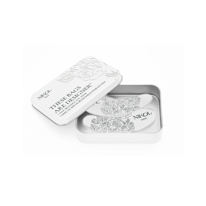 A rectangular metal tin with floral designs contains a pair of reusable, medical-grade silicone These Bags Are Designer-Eye Masks from Nikol Beauty. The partially open lid and masks feature floral patterns and the brand name.