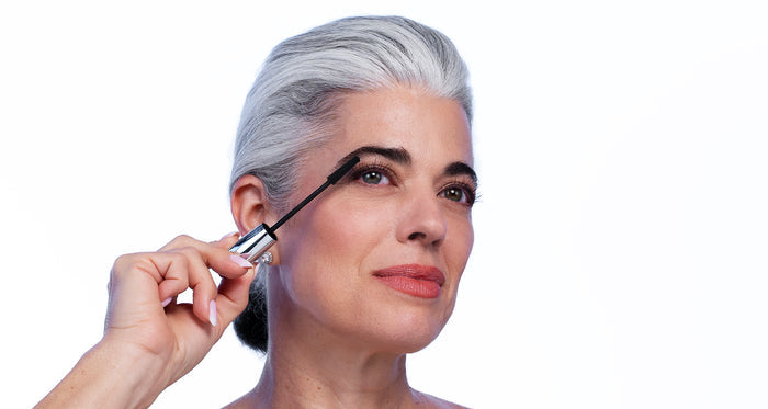 A woman with gray hair and light skin applies Nikol Beauty's Volume Up Mascara Bundle to her upper lashes, looking upward with a slight smile against a white background.
